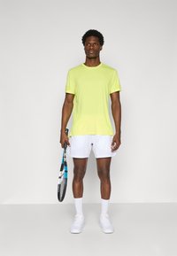 Lyhythihainen, kirkkaan keltainen urheilupaita pyöreällä kaula-aukolla, yhdistetty valkoisiin shortseihin. Malli pitää kädessään tennis mailaa ja käyttää valkoisia tennareita.