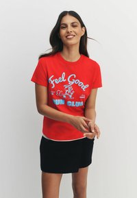 Sarkans kokvilnas T-krekls ar zilu grafisko uzrakstu "Feel Good Run Club", kombinēts ar melnu svārku, ar īsām piedurknēm.