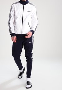Person som bär en vit och svart Adidas träningsjacka, svarta Adidas träningsbyxor, vita strumpor och svarta Adidas slidesandaler.
