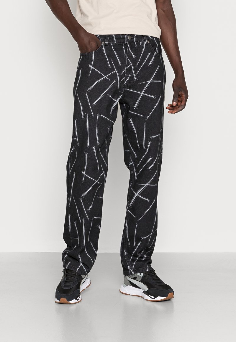 Pantalones de mezclilla negros con un patrón lineal blanco, que cuentan con un corte clásico, cinco bolsillos y un cierre de botón. Llevados con zapatillas negras.
