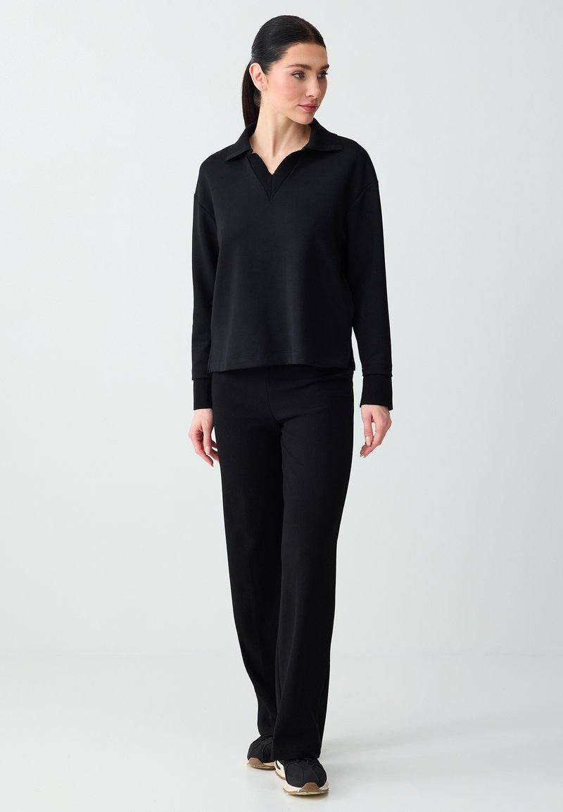 Femme debout, portant un haut noir à manches longues avec col, un pantalon noir et des baskets noires à semelles blanches, sur un fond clair uni.