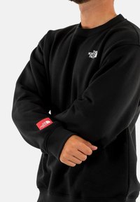 Sudadera negra de tejido suave con cuello redondo, puños acanalados y dos logotipos: uno blanco en el pecho y otro rojo en la manga.