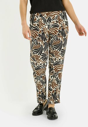 Femme portant un pantalon à motifs abstraits noirs, bruns et blancs, avec des mocassins noirs et un haut noir, se tenant devant un fond uni.