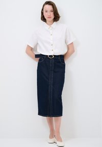 Blusa blanca de cuello con mangas cortas y detalles de botones dorados; falda lápiz de mezclilla oscura con cinturón y hebilla dorada. Zapatos planos blancos.