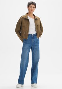 Braune kurze Jacke mit Reißverschluss, zwei vorderen Taschen; kombiniert mit hellblauen, hoch taillierten, weiten Jeans und weißen Sneakern.