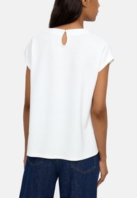 Biała bluzka z krótkim rękawem, okrągłym dekoltem i małym rozcięciem z tyłu. Gładka tekstura; lekko luźny krój. Połączona z ciemnoniebieskimi spodniami.