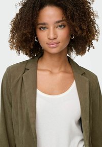 Olivgrüner Cord-Blazer mit strukturierter Stoffoberfläche, Reverskragen und lässiger Passform, über einem weißen Tanktop getragen. Kreisförmige silberne Ohrringe.