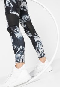 Czarno-białe, dopasowane legginsy z abstrakcyjnymi wzorami i siateczkowymi akcentami. W zestawieniu z białymi butami sportowymi, które podkreślają elegancki design.
