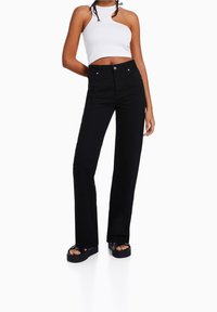 Pantalones de mezclilla anchos de mezclilla negra con cintura alta, que presentan bolsillos clásicos y cierre de botón. Combinados con un top corto de canalé blanco y sandalias de plataforma.