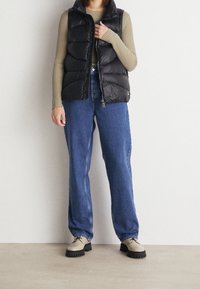 Gilet imbottito nero sopra un maglione beige a costine, abbinato a jeans a gamba larga blu e scarpe beige con suola testurizzata e dettagli di lacci.