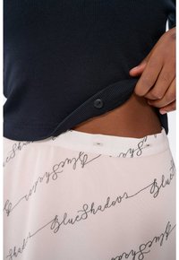 Schwarzes Rippenshirt mit einem dekorativen Knopf am Saum, kombiniert mit einem leichten Rock aus Stoff, der ein fließendes Schriftmuster mit der Aufschrift "Blaue Schatten" zeigt.