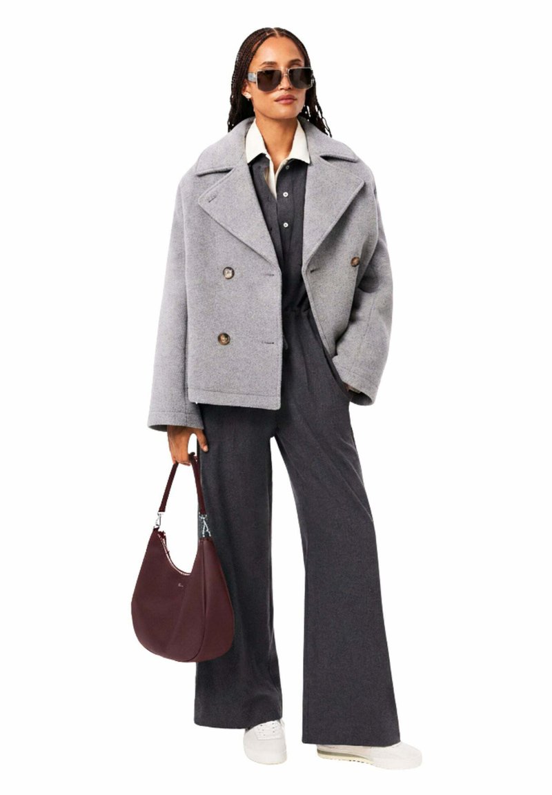 Frau trägt einen grauen, doppelreihigen Mantel, dunkelgraue Hosen mit weitem Bein, weiße Sneakers, Sonnenbrille und trägt eine dunkelrote Handtasche.