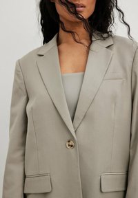 Light gray blazer with notched lapels, single button closure, front pockets, and a smooth texture. Worn over a matching top.