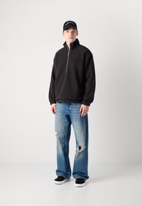 Tânăr îmbrăcat cu un hanorac din fleece negru cu fermoar pe jumătate, blugi rupți de culoare albastră, adidași negri și albi, și o șapcă neagră Calvin Klein, stând în fața unui fundal alb.