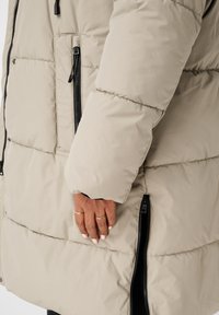 Doudoune beige avec un design matelassé, équipée d'une poche latérale avec une fermeture éclair noire et une texture légèrement brillante. Conçue pour la chaleur.