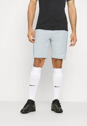 Homme portant un short Nike gris clair, des chaussettes blanches Nike hautes jusqu’aux genoux, des chaussures de sport Nike noires et un t-shirt noir sur un fond uni.