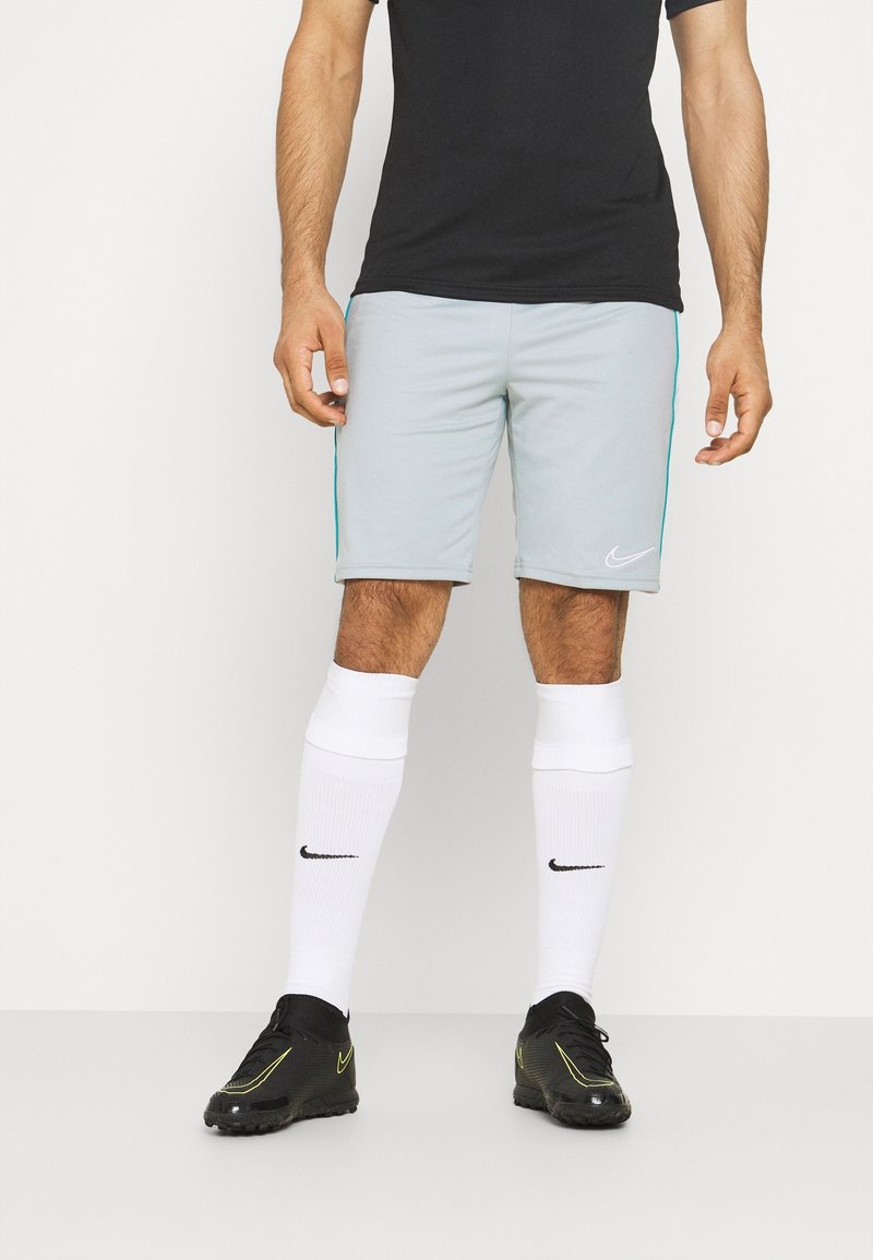 Homme portant un short Nike gris clair, des chaussettes blanches Nike hautes jusqu’aux genoux, des chaussures de sport Nike noires et un t-shirt noir sur un fond uni.