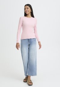 Sieviete, tērpusies gaiši rozā garām piedurknēm, plaša griezuma gaiši zilu džinsu biksēs un tan krāsas sandālēs, stāv un smaida pret baltu fona.