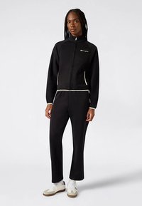 Zwarte atletische trainingspak met een jas met rits en bijpassende broek, voorzien van witte accentstiksels en een logo op de borst. Zacht materiaal, nauwsluitend ontwerp.