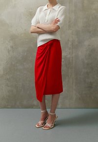 Witte gebreide polo met een gestructureerd patroon en korte mouwen, gecombineerd met een rode gedrapeerde midi-jurk. Beige sandalen met hoge hakken en banden.