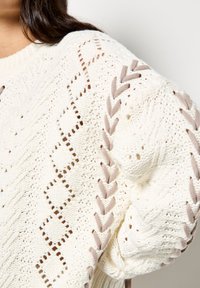 Gruby, kremowy sweter wykonany na szydełku, z otwartymi wzorami diametralnymi i jasnobrązowymi plecionymi akcentami na rękawach.