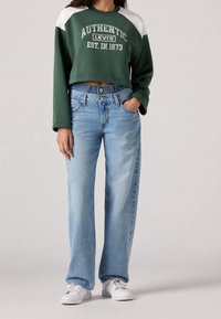 Persona vistiendo un sudadera corta de Levi's en color verde, jeans de cintura alta en azul claro y zapatillas blancas, de pie con una mano en el bolsillo.