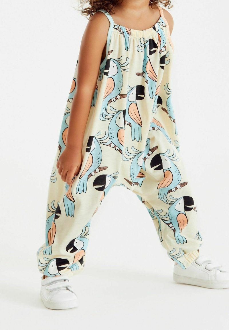 Combinaison jaune pâle avec un motif d'oiseaux colorés, encolure et poignets élastiqués, en tissu doux, associée à des baskets blanches.