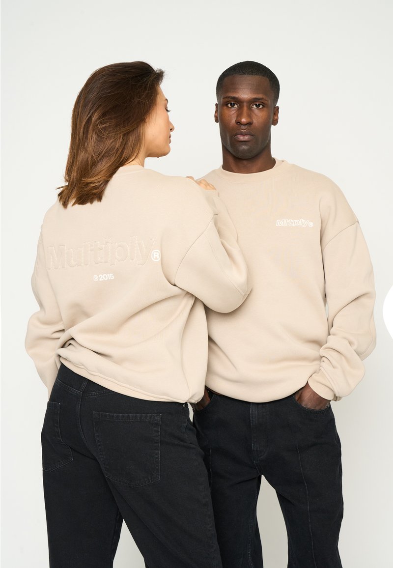 Beige Sweatshirts mit "Multiply" Logo, ausgestattet mit gerippten Bündchen und lockerer Passform, kombiniert mit schwarzen Jeans. Glatte Textur und lässiges Design.