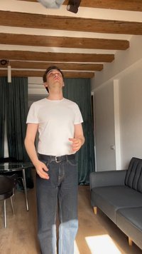 Un homme portant un t-shirt blanc et un jean foncé se tient à l'intérieur, lançant ou attrapant un petit objet gris au-dessus de sa tête.