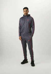 Gray athletic tracksuit with pink accents, zip-up hoodie, and tapered pants. Fabric appears lightweight and smooth. Black sneakers.