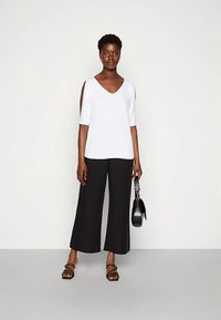 Weißes V-Ausschnitt-Oberteil mit ausgeschnittenen Ärmeln, kombiniert mit schwarzen, weit geschnittenen Hosen. Das Modell hält eine schwarze Handtasche und trägt schwarze Riemchensandalen.