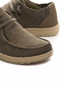 Zapatos de lona marrones con tejido texturizado, punta redonda y suela beige flexible. Presentan cordones atados y un sutil logotipo de la marca en el costado.