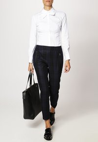 Biała koszula z długim rękawem i kołnierzykiem w kształcie muszki, zestawiona z granatowymi spodniami w kratę o dopasowanym kroju. Trzyma czarną torbę i ma na sobie czarne buty.