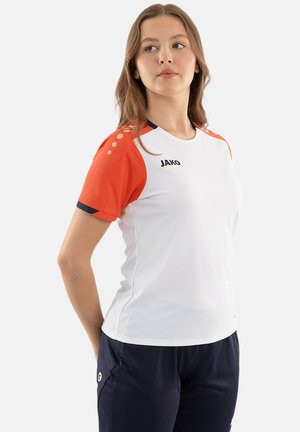 Giovane donna con indosso una maglietta sportiva bianca con maniche rosse e pantaloni blu navy, in piedi con le mani dietro la schiena, guardando di lato.