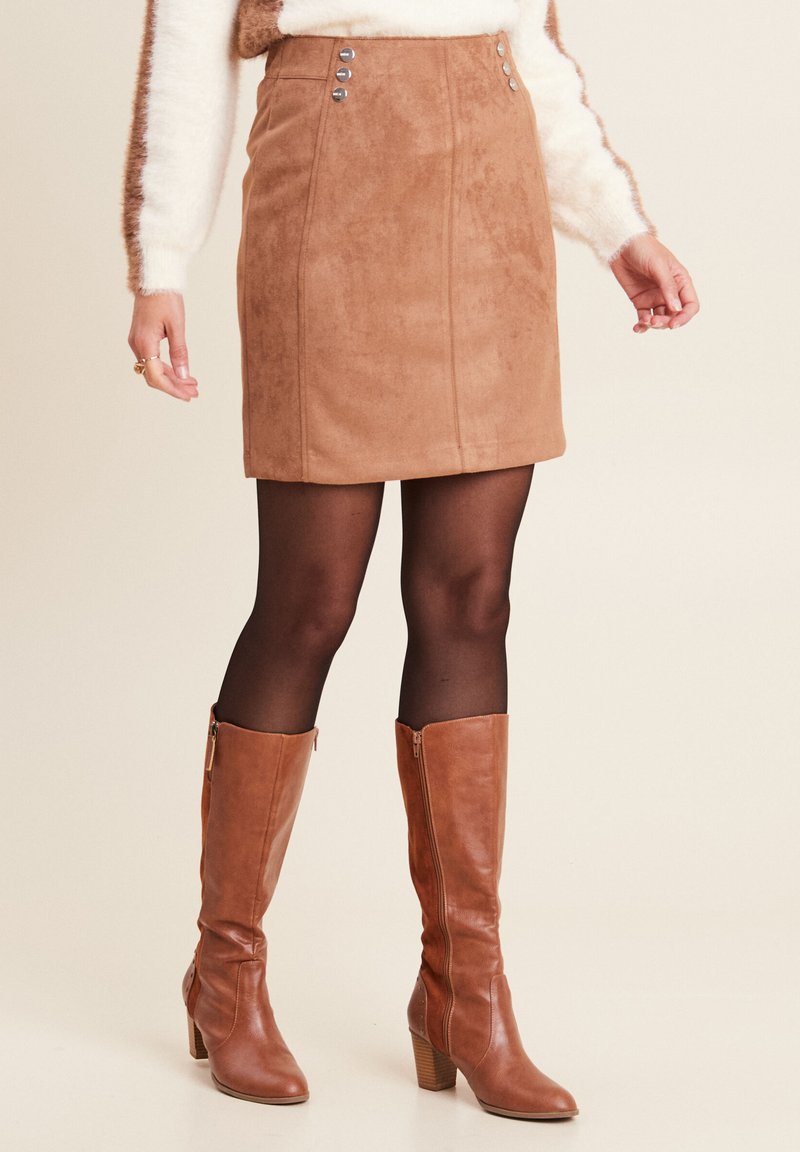 Frau, die einen braunen Wildlederrock, schwarze Strumpfhosen und passende knielange braune Lederstiefel vor einem schlichten hellen Hintergrund trägt.