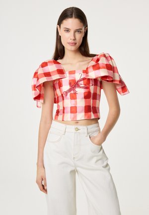 Woman wearing red and pink checkered crop top with puffed sleeves and lobster embroidery, paired with high-waisted white pants.