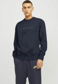 Marine crewneck sweatshirt met verhoogde "ORIGINALS" tekst, geribbelde boorden en zoom. Gepaard met grijze sweatpants met gele logo.