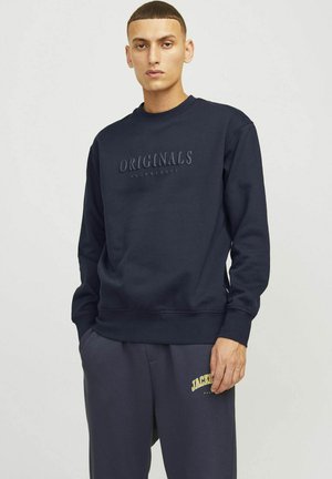 Sudadera de cuello redondo en color marino con el texto grabado "ORIGINALS", puños y dobladillo acanalados. Combinada con pantalones de chándal grises que presentan un logo amarillo.