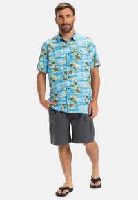Blå kortærmet skjorte med et mønster af palmer og strand, parret med grå cargo shorts og sorte flip-flops. Modellen står selvsikkert.