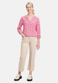 Rosa gemusterte Bluse mit V-Ausschnitt, gerafften Ärmeln und Knopfdetails, kombiniert mit beigen, cropped Hosen und weißen Fransen-Loafern.