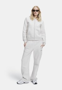 Lys grå hettegenser med glidelås og frontlommer, matchende joggebukser, med en avslappet passform og en subtil logo på venstre side.