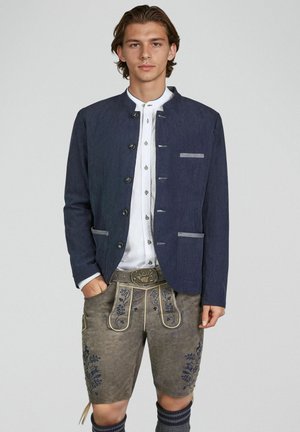 Mann trägt traditionelle bayerische Tracht mit marineblauer Jacke, weißem Hemd, bestickter Lederhose und einem dekorativen Gürtel.