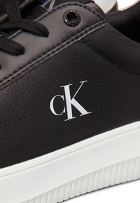 Schwarze Sneaker aus strukturiertem Synthetikleder, mit einem weißen Logo, schwarzen Schnürsenkeln und einer weißen Gummisohle mit Rillen.