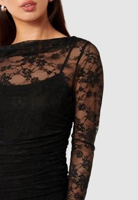 Μαύρο φόρεμα δαντέλας με μακριά διάφανα μανίκια και εφαρμοστό κορσέ με μαζεμένη υφή. Σχεδιασμένο με λαιμόκοψη μπαούλας και πολύπλοκα λουλουδάτα σχέδια.