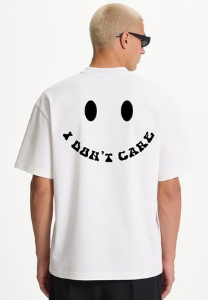 Person with short blond hair wearing black sunglasses and a white t-shirt with a black smiley face and "I DON'T CARE" text on the back.
