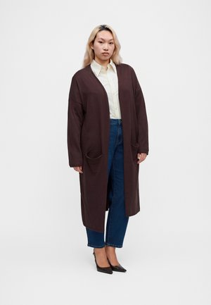 Jeune femme aux cheveux clairs portant un long manteau marron foncé, une chemise blanche boutonnée, un jean bleu et des chaussures noires pointues, debout sur un fond blanc.