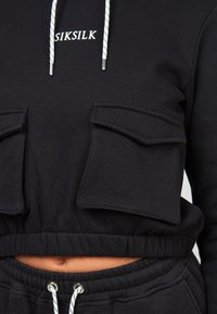 Felpa nera corta con due tasche frontali, cappuccio con cordino e cintura elastica. Presenta ricamo del logo e tessuto testurizzato.