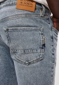 Τα τζιν ελαφριάς απόχρωσης διαθέτουν πίσω τσέπη με αντίθετη ραφή και δερμάτινη ετικέτα που αναγράφει "Scotch & Soda Amsterdam."