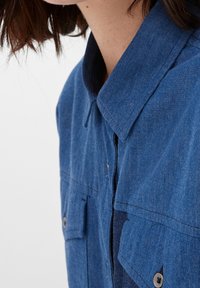 Kvinde iført en blå denimskjorte med knappede brystlommer, synlig krave og brunt hår, der delvist dækker nakkeregionen.