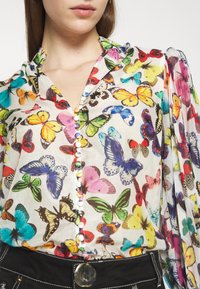 Gros plan d'une personne portant un chemisier imprimé papillons colorés avec des boutons multicolores recouverts et un pantalon noir avec des coutures blanches.